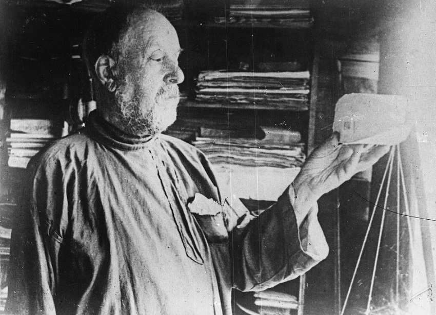 К. Э. Циолковский в своей мастерской с бумажной моделью в руке. 1932.  Фотограф: Чмиль Феодосий Андроникович (1900-1945). Цифровая копия оригинальной черно-белой фотографии Константина Эдуардовича Циолковского. Источник: Wikimedia Commons (Public Domain).